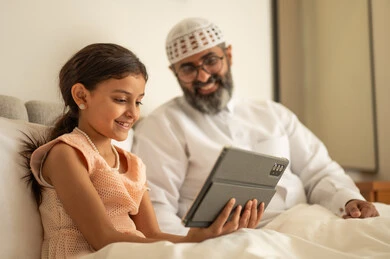 Integrating learning with technology and tech, following available online e-learning platforms, watching recreational and educational activities, an Arab Gulf Saudi girl wearing a dress sitting on the bed with her father using a tablet, gestures of happiness and joy.