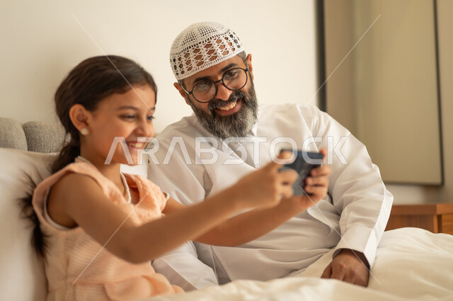 اب عربي خليجي سعودي يرتدي الثوب التقليدي والقبعة البيضاء يجلس على السرير مع ابنته، فتاة عربية خليجية سعودية تحمل بيدها الهاتف المحمول، استخدام جهاز تقني حديث ومتطور، مفهوم التعليم الالكتروني، متابعة الدروس عن بعد عن طريق الانترنت، المناهج العلمية التعليمية