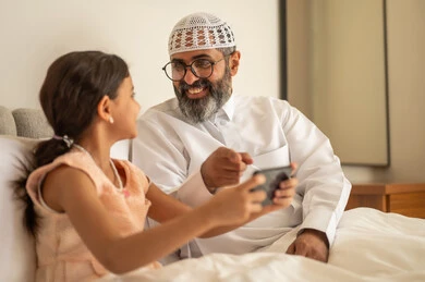 Using a modern and advanced technical device, an Arab Gulf Saudi father wearing traditional attire and a white cap sits on the bed with his daughter, a Gulf Arab Saudi girl holding a mobile phone in her hand, the concept of e-learning, following lessons remotely via the internet, educational scientific curricula.