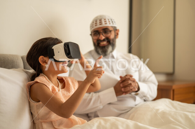 الاشارة الى شيء ما, فتاة عربية خليجية سعودية تضع نظارة الواقع الافتراضي, المتعة والتسلية بالمشاهدة عبر نظارات ثلاثية الابعاد، التفاعل والاندماج ومحاكاة العالم الخيالي، رجل عربي خليجي سعودي مبتسم يرتدي الثوب التقليدي والقبعة البيضاء يجلس على السرير مع ابنته