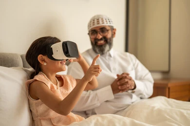 الاشارة الى شيء ما, فتاة عربية خليجية سعودية تضع نظارة الواقع الافتراضي, المتعة والتسلية بالمشاهدة عبر نظارات ثلاثية الابعاد، التفاعل والاندماج ومحاكاة العالم الخيالي، رجل عربي خليجي سعودي مبتسم يرتدي الثوب التقليدي والقبعة البيضاء يجلس على السرير مع ابنته