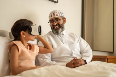 A Saudi Arabian Gulf girl is wearing virtual reality glasses, enjoying and entertaining herself by watching through 3D glasses, interacting and immersing herself in a simulated fantasy world. A smiling Saudi Arabian Gulf man, dressed in traditional attire and a white headscarf, is sitting on the bed with his daughter.