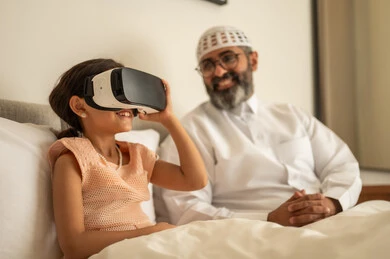 Enjoyment and entertainment through watching with 3D glasses, interaction and immersion simulating a fantasy world, a smiling Arab Gulf Saudi man wearing traditional attire and a white cap sitting on the bed with his daughter, an Arab Gulf Saudi girl wearing virtual reality glasses.