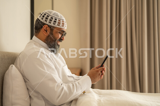 Browsing social media, an Arab Gulf Saudi man wearing traditional attire and a white cap sits on the bed holding a mobile phone, chatting and communicating with family and friends, using a modern and advanced technological device, working remotely and completing the required tasks.