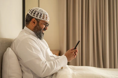تصفح مواقع التواصل الاجتماعي، رجل عربي خليجي سعودي يرتدي الثوب التقليدي والقبعة البيضاء يجلس على السرير يمسك بيده الهاتف المحمول، الدردشة والتواصل مع الأهل و الأصدقاء، استخدام جهاز تقني حديث ومتطور، العمل عن بعد وانجاز المهام المطلوبة