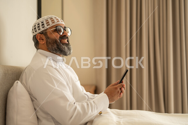النظر الى شيء ما, تصفح مواقع التواصل الاجتماعي، رجل عربي خليجي سعودي يرتدي الثوب التقليدي والقبعة البيضاء يجلس على السرير يمسك بيده الهاتف المحمول، الدردشة والتواصل مع الأهل و الأصدقاء، استخدام جهاز تقني حديث ومتطور، العمل عن بعد وانجاز المهام المطلوبة