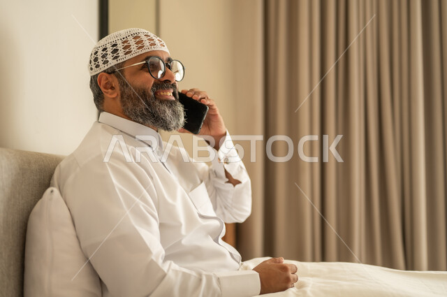 اجراء مكالمة هاتفية عبر الجوال، رجل عربي خليجي سعودي يرتدي الثوب التقليدي والقبعة البيضاء يجلس على السرير يمسك بيده الهاتف المحمول، الدردشة والتواصل مع الأهل و الأصدقاء، استخدام جهاز تقني حديث ومتطور