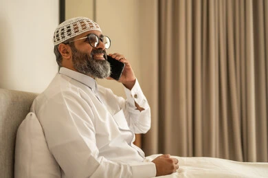 اجراء مكالمة هاتفية عبر الجوال، رجل عربي خليجي سعودي يرتدي الثوب التقليدي والقبعة البيضاء يجلس على السرير يمسك بيده الهاتف المحمول، الدردشة والتواصل مع الأهل و الأصدقاء، استخدام جهاز تقني حديث ومتطور