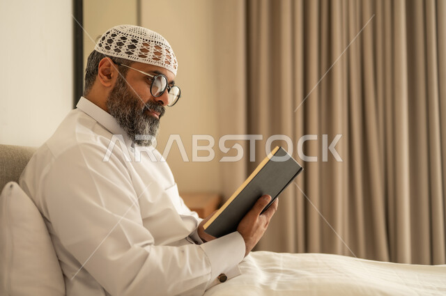تنمية مهارة القراءة, قضاء أوقات الفراغ بالتثقيف والتعلم, الاهتمام بتصفح الكتب المفيدة، رجل عربي خليجي سعودي يرتدي الثوب التقليدي والقبعة البيضاء يجلس على السرير في المنزل ويحمل بيده كتاب