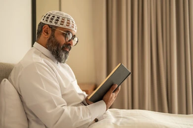 تنمية مهارة القراءة, قضاء أوقات الفراغ بالتثقيف والتعلم, الاهتمام بتصفح الكتب المفيدة، رجل عربي خليجي سعودي يرتدي الثوب التقليدي والقبعة البيضاء يجلس على السرير في المنزل ويحمل بيده كتاب