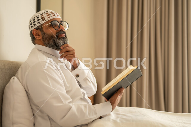 Gestures of thinking about something, developing reading skills, spending free time on education and learning, being interested in browsing useful books, an Arab Gulf Saudi man wearing traditional attire and a white cap sitting on the bed at home holding a book in his hand.
