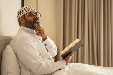 ايماءات التفكير فى شيء ما, تنمية مهارة القراءة, قضاء أوقات الفراغ بالتثقيف والتعلم, الاهتمام بتصفح الكتب المفيدة، رجل عربي خليجي سعودي يرتدي الثوب التقليدي والقبعة البيضاء يجلس على السرير في المنزل ويحمل بيده كتاب