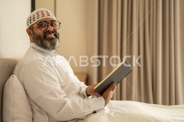 قضاء أوقات الفراغ بالتثقيف والتعلم, تنمية مهارات القراءة, الاهتمام بتصفح الكتب المفيدة، رجل عربي خليجي سعودي يرتدي الثوب التقليدي والقبعة البيضاء يجلس على السرير في المنزل ويحمل بيده كتاب