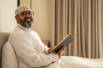Spending free time on education and learning, developing reading skills, and being interested in browsing useful books. An Arab Gulf Saudi man wearing traditional attire and a white cap is sitting on the bed at home holding a book in his hand.