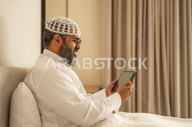 إدارة شؤون الأعمال وتنظيمها عبر الاجهزة التقنية، مهنة ووظيفة ادارية، دمج العمل مع التكنولوجيا، رجل عربي خليجي سعودي يرتدي الثوب التقليدي والقبعة البيضاء يجلس على السرير يعمل على الجهاز اللوحي، استخدام جهاز تقني حديث ومتطور، العمل عن بعد وانجاز المهام المطلوبة