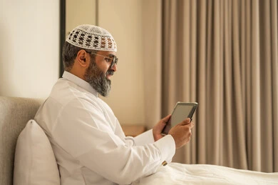 إدارة شؤون الأعمال وتنظيمها عبر الاجهزة التقنية، مهنة ووظيفة ادارية، دمج العمل مع التكنولوجيا، رجل عربي خليجي سعودي يرتدي الثوب التقليدي والقبعة البيضاء يجلس على السرير يعمل على الجهاز اللوحي، استخدام جهاز تقني حديث ومتطور، العمل عن بعد وانجاز المهام المطلوبة