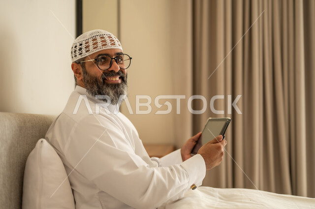 النظر الى الكاميرا بتعابير السرور, إدارة شؤون الأعمال وتنظيمها عبر الاجهزة التقنية، مهنة ووظيفة ادارية، دمج العمل مع التكنولوجيا، رجل عربي خليجي سعودي يرتدي الثوب التقليدي والقبعة البيضاء يجلس على السرير يعمل على الجهاز اللوحي، استخدام جهاز تقني حديث ومتطور، العمل عن بعد وانجاز المهام المطلوبة