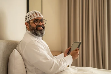 النظر الى الكاميرا بتعابير السرور, إدارة شؤون الأعمال وتنظيمها عبر الاجهزة التقنية، مهنة ووظيفة ادارية، دمج العمل مع التكنولوجيا، رجل عربي خليجي سعودي يرتدي الثوب التقليدي والقبعة البيضاء يجلس على السرير يعمل على الجهاز اللوحي، استخدام جهاز تقني حديث ومتطور، العمل عن بعد وانجاز المهام المطلوبة