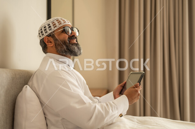 النظر الى شيء ما, إدارة شؤون الأعمال وتنظيمها عبر الاجهزة التقنية، مهنة ووظيفة ادارية، دمج العمل مع التكنولوجيا، رجل عربي خليجي سعودي يرتدي الثوب التقليدي والقبعة البيضاء يجلس على السرير يعمل على الجهاز اللوحي، استخدام جهاز تقني حديث ومتطور، العمل عن بعد وانجاز المهام المطلوبة