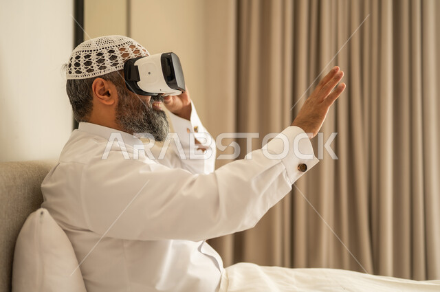 رفع كف اليد للأعلى بإيماءات الاندماج ومحاكاة العالم الخيالي، رجل عربي خليجي سعودي مبتسم يرتدي الثوب التقليدي والقبعة البيضاء بجلس على السرير يضع نظارة الواقع الافتراضي، المتعة والتسلية بالمشاهدة عبر نظارات ثلاثية الابعاد