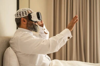 رفع كف اليد للأعلى بإيماءات الاندماج ومحاكاة العالم الخيالي، رجل عربي خليجي سعودي مبتسم يرتدي الثوب التقليدي والقبعة البيضاء بجلس على السرير يضع نظارة الواقع الافتراضي، المتعة والتسلية بالمشاهدة عبر نظارات ثلاثية الابعاد