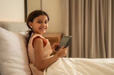 متابعة الدروس عن بعد عن طريق الانترنت, مفهوم التعليم الالكتروني، المناهج العلمية التعليمية، فتاة عربية خليجية سعودية ترتدي الفستان تجلس على السرير تحمل بيدها الجهاز اللوحي، استخدام جهاز تقني حديث ومتطور