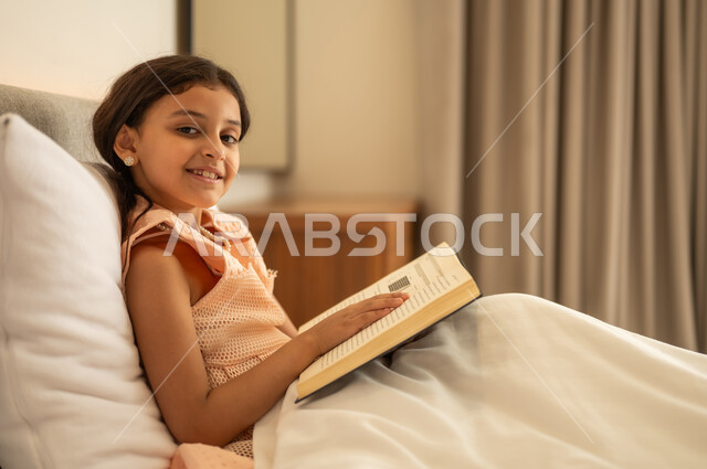 النظر للكاميرا بتعابير السرور, الاهتمام بتصفح الكتب المفيدة, تنمية مهارات القراءة للأطفال, تمضية أوقات الفراغ بالتعلم والتثقف الذاتي, فتاة عربية خليجية سعودية تجلس على السرير تقوم بالقراءة بتمعن, مطالعة القصص التصويرية المسلية