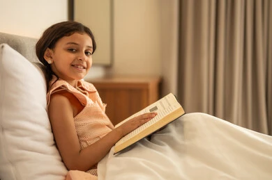 Looking at the camera with expressions of joy, taking an interest in browsing useful books, developing reading skills for children, spending free time learning and self-educating, an Arab Gulf Saudi girl sitting on the bed reading attentively, exploring entertaining picture stories.