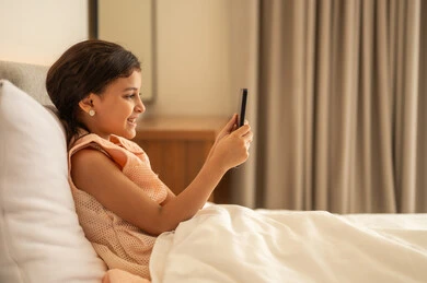 Exchanging conversations and news, communicating and chatting with family and friends, browsing social media applications and programs, a Gulf Arab Saudi girl wearing a dress sitting on the bed holding a mobile phone in her hand, using modern and advanced technological devices.
