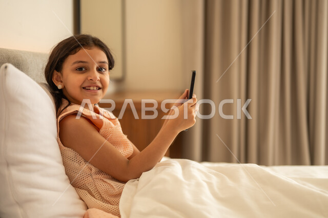 Communication and chatting with family and friends, browsing social media applications and programs, exchanging conversations and news, a Gulf Arab Saudi girl wearing a dress sitting on the bed holding a mobile phone in her hand, using modern and advanced technological devices.