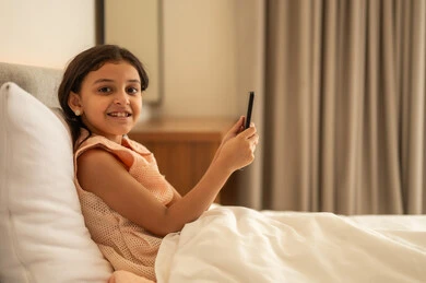 التواصل والدردشه مع الأهل والأصدقاء، تصفح تطبيقات وبرامج التواصل الاجتماعي، تبادل الاحاديث والاخبار، فتاة عربية خليجية سعودية ترتدي فستان تجلس على السرير تمسك بيدها الهاتف المحمول، استخدام الاجهزة التقنية الحديثة والمتطورة