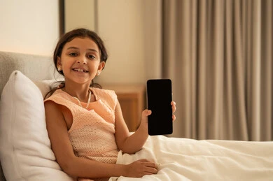 عرض شاشة سوداء فارغة، التواصل والدردشه مع الأهل والأصدقاء، تصفح تطبيقات وبرامج التواصل الاجتماعي، تبادل الاحاديث والاخبار، فتاة عربية خليجية سعودية ترتدي فستان تجلس على السرير تمسك بيدها الهاتف المحمول، استخدام الاجهزة التقنية الحديثة والمتطورة