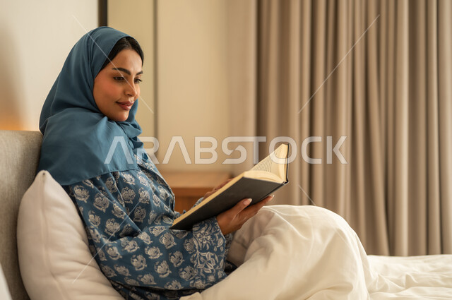 تمضية أوقات الفراغ بالتعلم والتثقف الذاتي, إيماءات الانتباه والتركيز بالمطالعة, امراة عربية خليجية سعودية محجبة ترتدي العباءة تجلس على السرير تقوم بالقراءة بتمعن قبل النوم , مطالعة القصص التصويرية المسلية, تنمية مهارات القراءة, الاهتمام بتصفح الكتب المفيدة