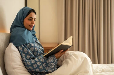 تمضية أوقات الفراغ بالتعلم والتثقف الذاتي, إيماءات الانتباه والتركيز بالمطالعة, امراة عربية خليجية سعودية محجبة ترتدي العباءة تجلس على السرير تقوم بالقراءة بتمعن قبل النوم , مطالعة القصص التصويرية المسلية, تنمية مهارات القراءة, الاهتمام بتصفح الكتب المفيدة