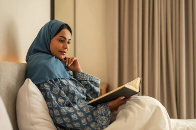 Spending leisure time learning and self-education, gestures of attention and focus through reading, a veiled Saudi Arabian woman wearing an abaya sits on the bed reading attentively before sleep, reading entertaining picture books, developing reading skills, and browsing useful books.