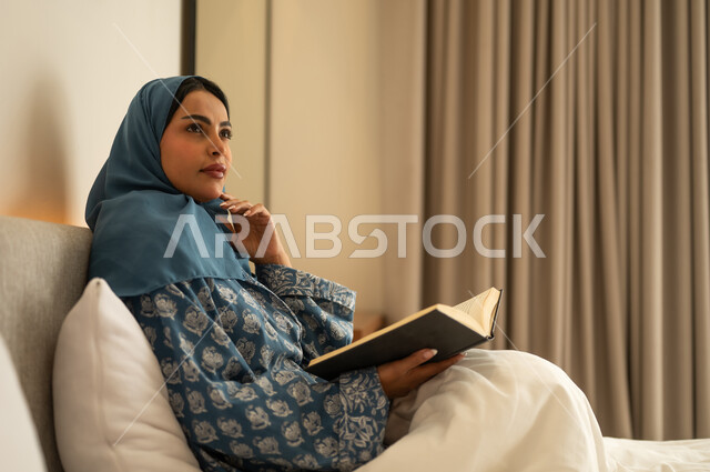 Looking at something, spending free time learning and self-education, a veiled Saudi Arabian woman wearing an abaya sits on the bed reading intently before sleep, reading entertaining picture books, developing reading skills, and browsing useful books.