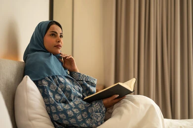 Looking at something, spending free time learning and self-education, a veiled Saudi Arabian woman wearing an abaya sits on the bed reading intently before sleep, reading entertaining picture books, developing reading skills, and browsing useful books.