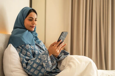 Chatting with family and friends, browsing social media apps and programs, working remotely, a smiling, veiled Saudi Arabian woman wearing an abaya, sitting on a bed holding her mobile phone, using modern and advanced technological devices.
