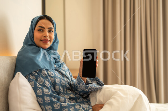 Displaying a blank black screen on a mobile phone, chatting with family and friends, browsing social media apps and programs, working remotely, a smiling, veiled Saudi Arabian woman wearing an abaya, sitting on a bed holding her mobile phone, using modern and advanced technological devices.