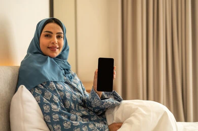 Displaying a blank black screen on a mobile phone, chatting with family and friends, browsing social media apps and programs, working remotely, a smiling, veiled Saudi Arabian woman wearing an abaya, sitting on a bed holding her mobile phone, using modern and advanced technological devices.