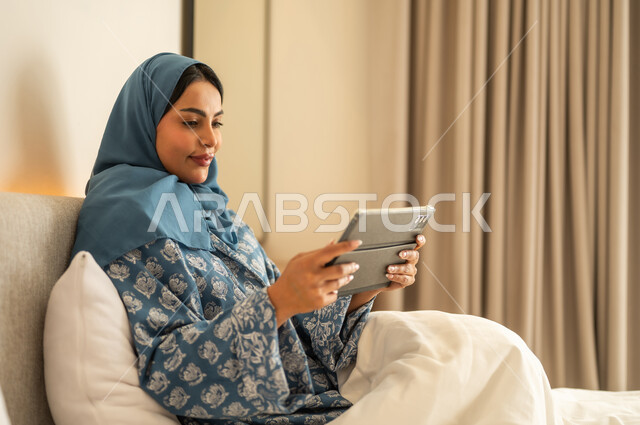 تصفح تطبيقات وبرامج التواصل الاجتماعي، الدردشه مع الأهل والأصدقاء، العمل عن بعد عبر الانترنت، امراة عربية خليجية سعودية محجبة مبتسمة ترتدي عباءة تجلس على السرير تمسك بيدها التابليت المحمول، استخدام الاجهزة التقنية الحديثة والمتطورة