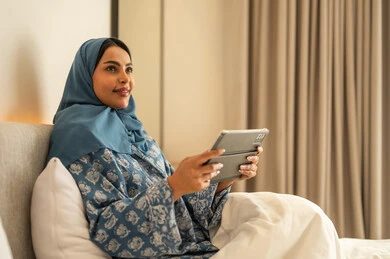 الدردشه مع الأهل والأصدقاء، العمل عن بعد عبر الانترنت، تصفح تطبيقات وبرامج التواصل الاجتماعي، امراة عربية خليجية سعودية محجبة مبتسمة ترتدي عباءة تجلس على السرير تمسك بيدها التابليت المحمول، استخدام الاجهزة التقنية الحديثة والمتطورة