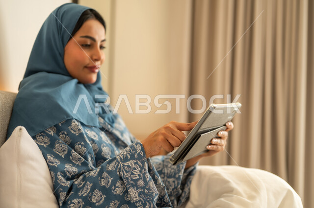 العمل عن بعد عبر الانترنت، الدردشه مع الأهل والأصدقاء، تصفح تطبيقات وبرامج التواصل الاجتماعي، امراة عربية خليجية سعودية محجبة مبتسمة ترتدي عباءة تجلس على السرير تمسك بيدها التابليت المحمول، استخدام الاجهزة التقنية الحديثة والمتطورة