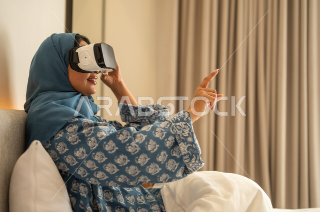 الاشارة الى شئ ما بإيماءات الاندماج ومحاكاة العالم الخيالي، قضاء وقت الفراغ بشكل ممتع، المتعة و التسلية بالمشاهدة عبر نظارة الواقع الافتراضي، إمرأة عربية خليجية سعودية محجبة ترتدي العباءة تجلس على السرير تنظر من خلال نظارات ثلاثية الأبعاد