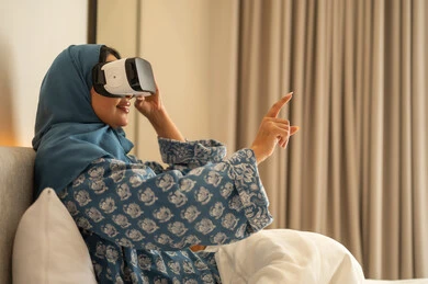 Referring to something through immersive gestures and simulating the imaginary world, spending leisure time in an enjoyable way, the fun and entertainment of watching through virtual reality glasses, a Saudi Arabian Gulf woman wearing a hijab and an abaya sitting on the bed looking through 3D glasses.