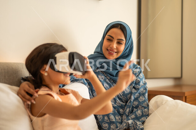 Trying to touch something, merging and simulating the imaginary world, the fun and entertainment of watching through virtual reality glasses, a Saudi Gulf Arab girl wearing a dress sitting on the bed with her mother and looking through 3D glasses, gestures of happiness and joy, technological and technical advancement.