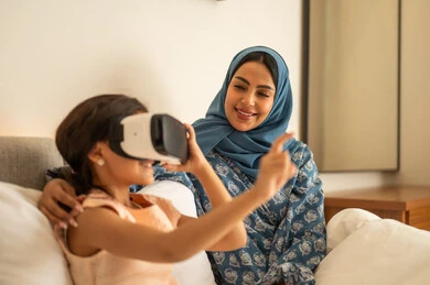 Trying to touch something, merging and simulating the imaginary world, the fun and entertainment of watching through virtual reality glasses, a Saudi Gulf Arab girl wearing a dress sitting on the bed with her mother and looking through 3D glasses, gestures of happiness and joy, technological and technical advancement.