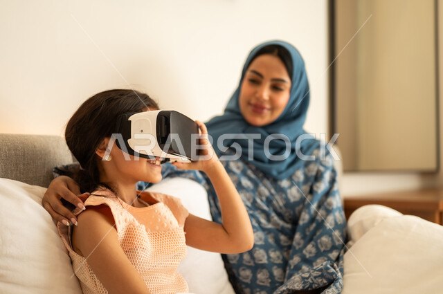 المتعة و التسلية بالمشاهدة عبر نظارة الواقع الافتراضي، فتاة عربية خليجية سعودية ترتدي الفستان تجلس على السرير مع والدتها وتنظر من خلال نظارات ثلاثية الأبعاد، الاندماج ومحاكاة العالم الخيالي ، ايماءات السعادة والسرور، التطور التقني والتكنولوجي
