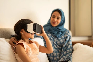 The enjoyment and entertainment of watching through virtual reality glasses, a Gulf Arab Saudi girl wearing a dress sitting on the bed with her mother and looking through 3D glasses, the immersion and simulation of the imaginary world, gestures of happiness and joy, technological and technical advancement.