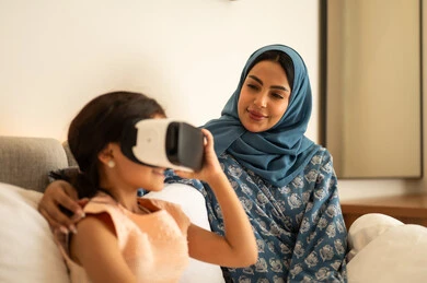 الاندماج ومحاكاة العالم الخيالي، المتعة و التسلية بالمشاهدة عبر نظارة الواقع الافتراضي، فتاة عربية خليجية سعودية ترتدي الفستان تجلس على السرير مع والدتها وتنظر من خلال نظارات ثلاثية الأبعاد، ايماءات السعادة والسرور، التطور التقني والتكنولوجي